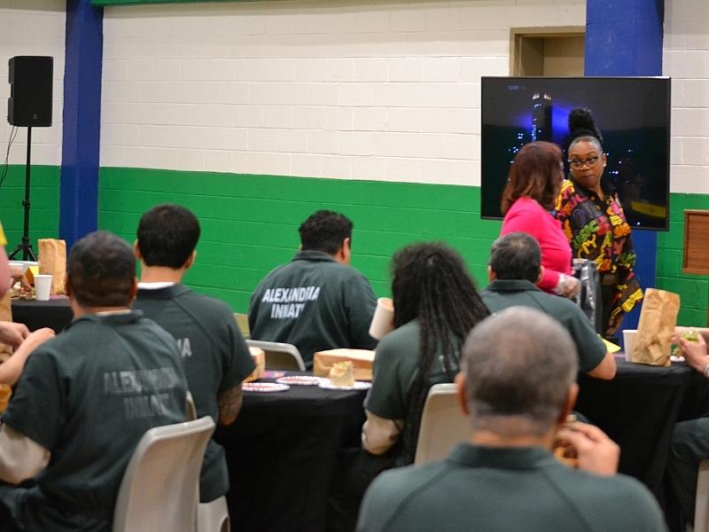 several inmates in green jumpsuits seated at table and eating with two civilian staff in background
