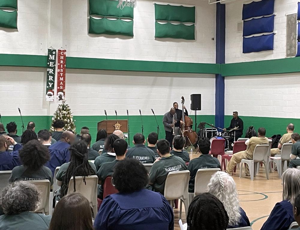 about 30 inmates seated in a gym with three musicians performing and Christmas decorations in the background