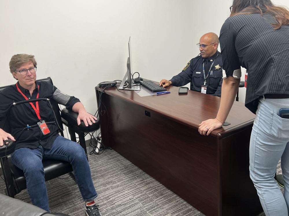 Civilan sitting in chair with polygraph equipment attached, deputy at desk looking at screen and another civilian leaning over desk and watching