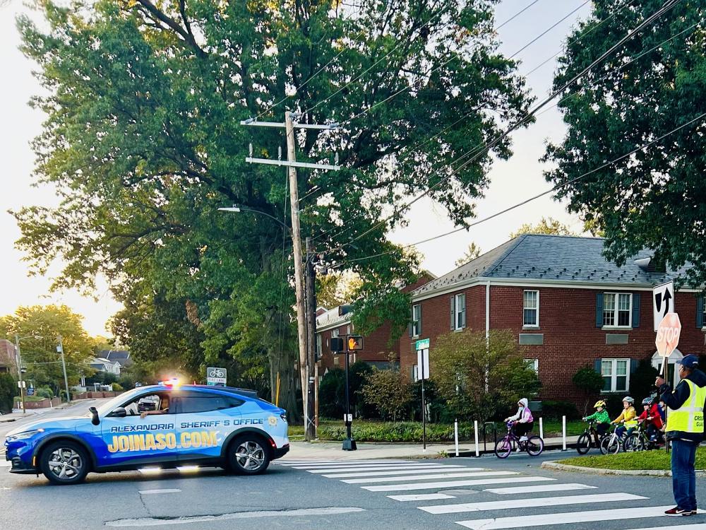 Blue and gold sheriff's cruiser with blue and red lights flashing escorting several children riding bicycles down a street, with a crossing guard visible on the right at a crosswalk