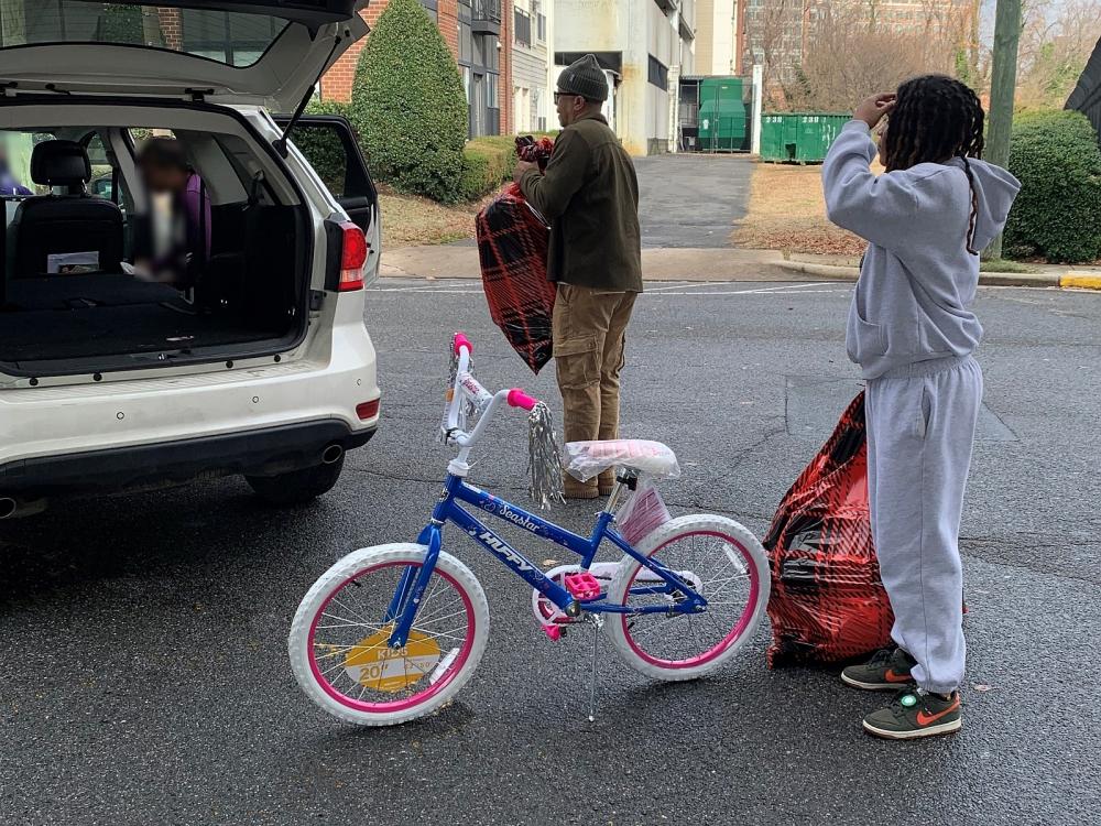 two civilian employees loading a child's bicycle and two large bags full of gifts into a car
