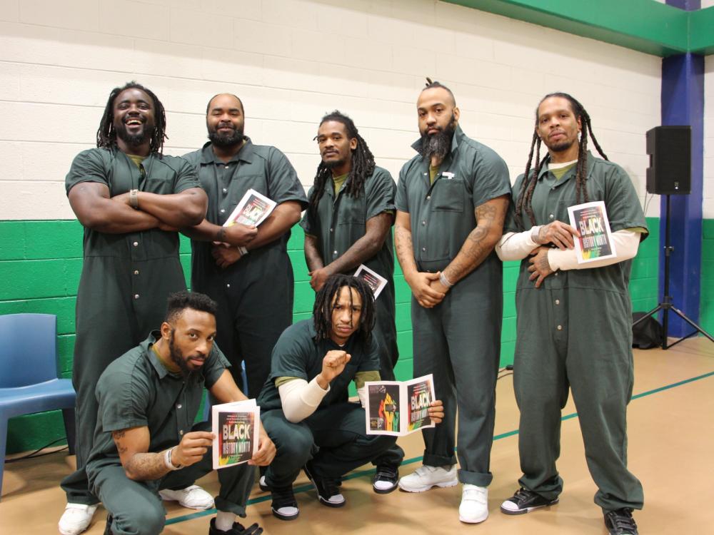 Seven inmates in green jumpsuits, with some holding printed programs for Black History Month,