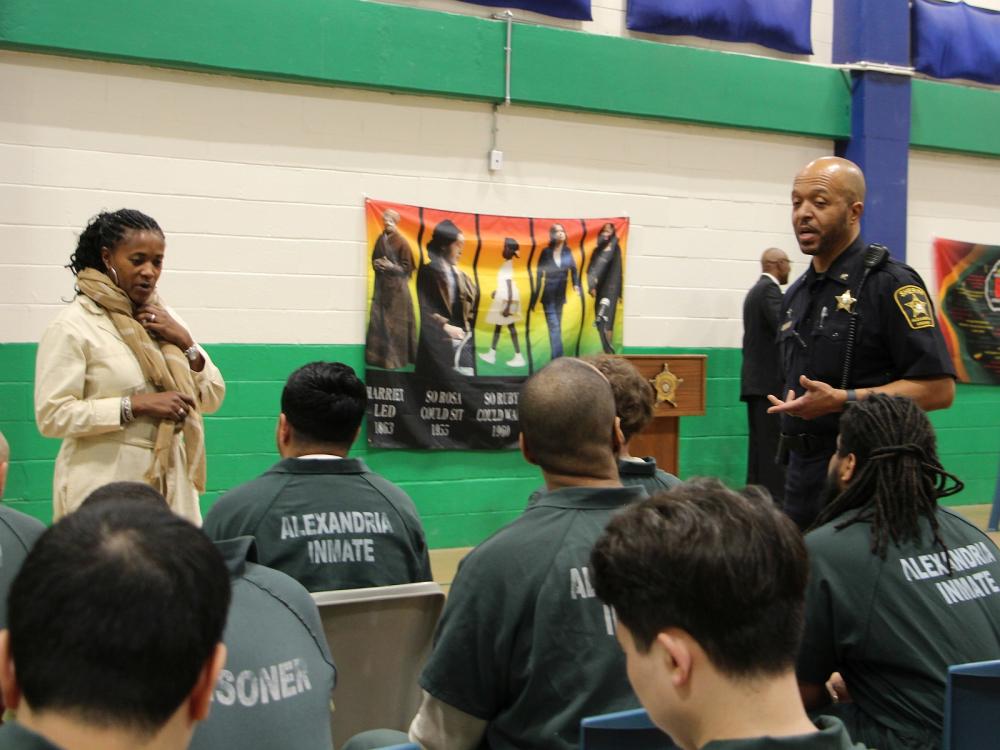 Professional staff member and deputy in blue uniform speak with audience members wearing green jumpsuits before the Black History Month program begins