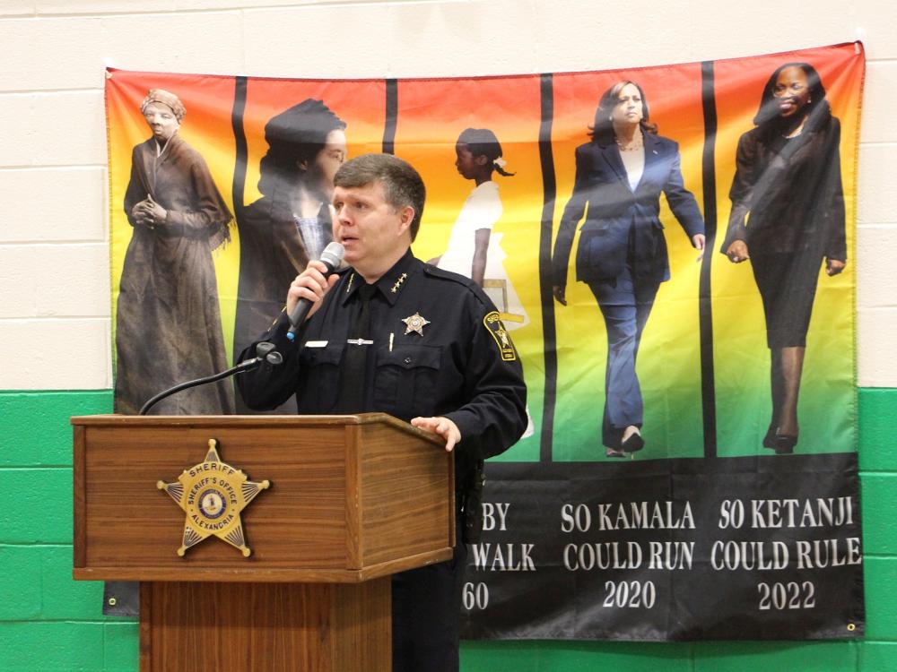Sheriff in blue uniform standing at podium and speaking into a microphone with a banner of Harriet Tubman, Rosa Parks, Ruby Bridges, Kamala Harris and Ketanji Brown Jackson in the background