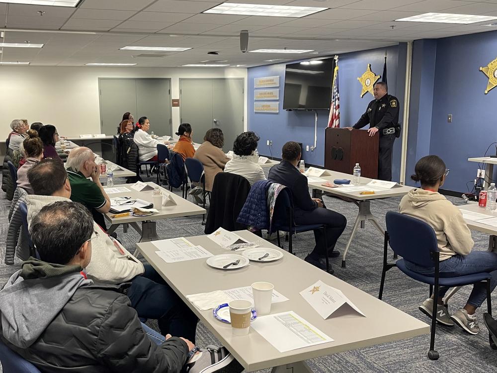 Sheriff's Office commander in blue uniform standing a podium and speaking to several community members in a classroom setting