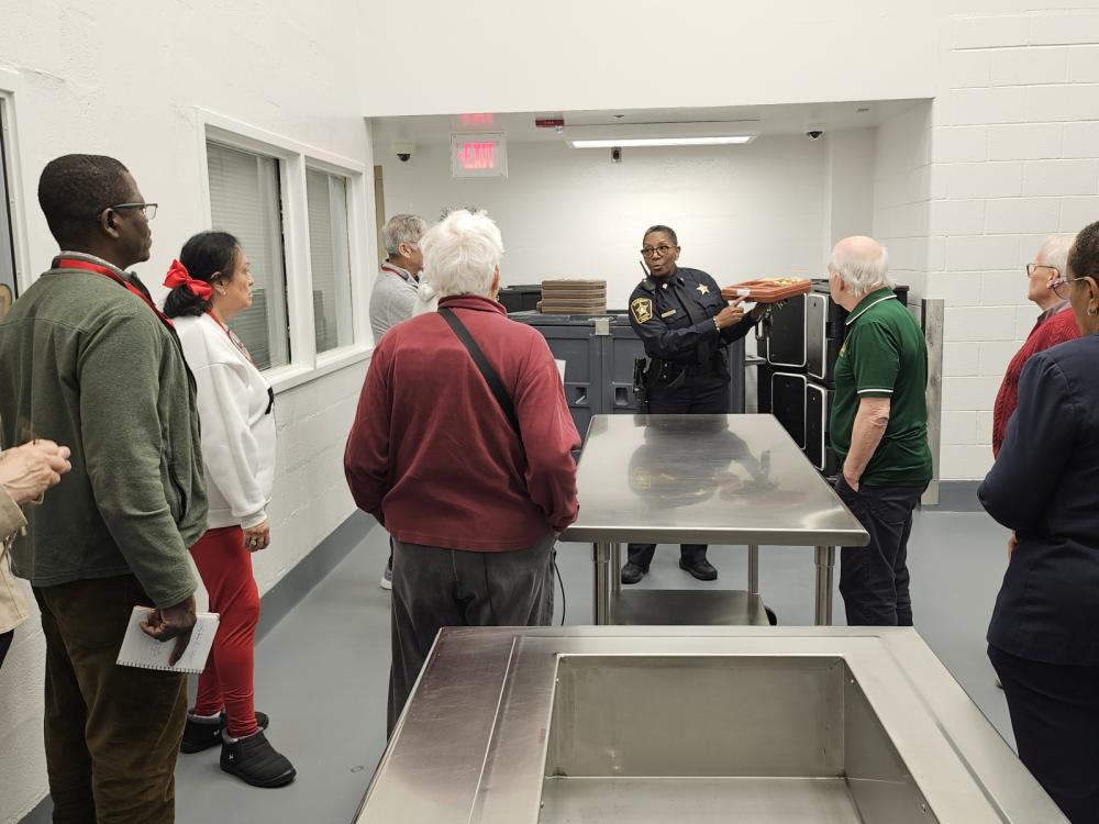 Deputy in blue uniform holding meal service tray of food inside institutional kitchen with several community members present