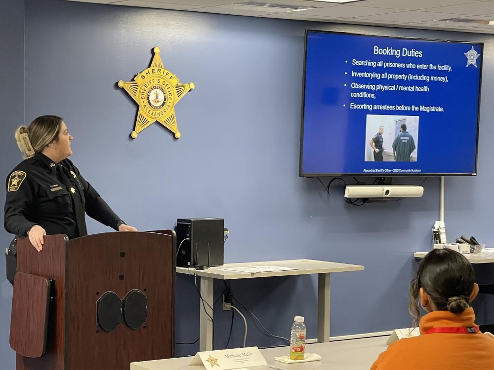 Sergeant in a blue uniform standing a podium and looking toward a screen for a presentation on the booking process