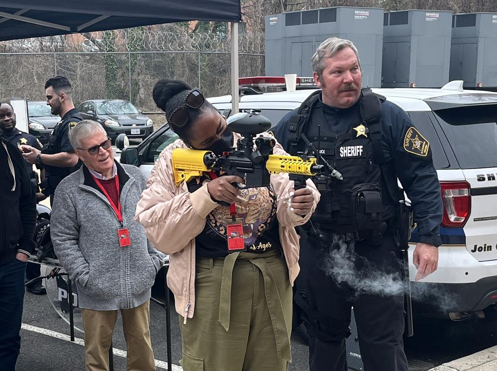 Civilian firing a pepperball training launcher under the supervision of a Sheriff's sergeant in a blue uniform.