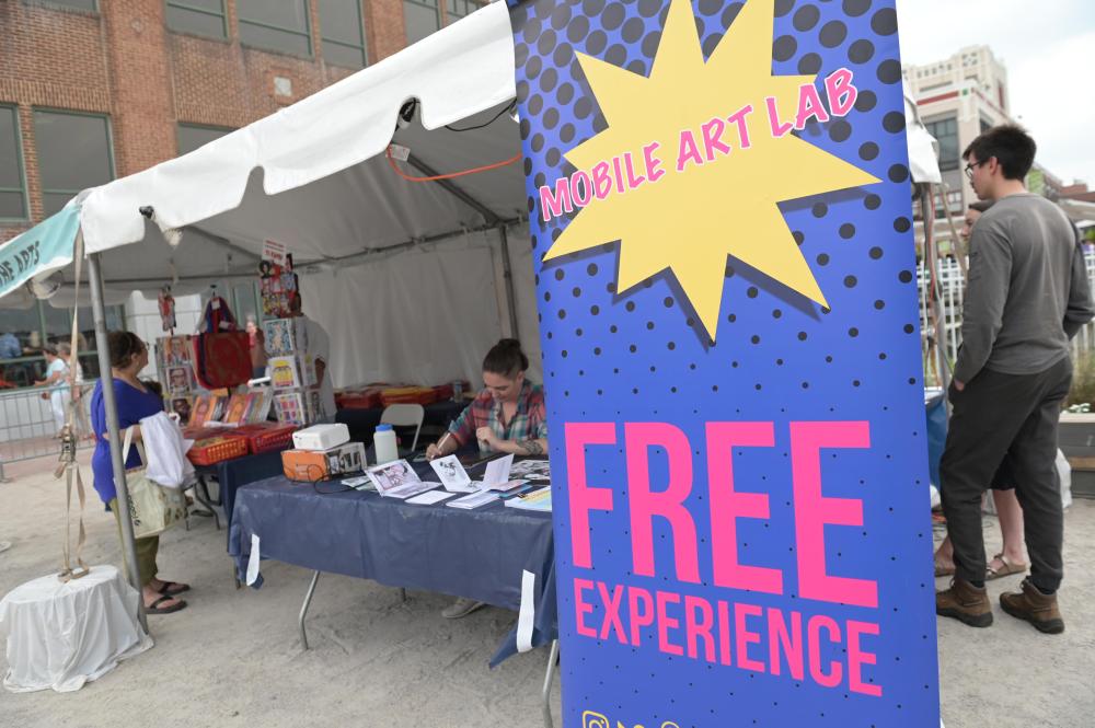  A vibrant event booth under a white canopy tent with a large blue and pink sign reading "Mobile Art Lab – FREE EXPERIENCE." 