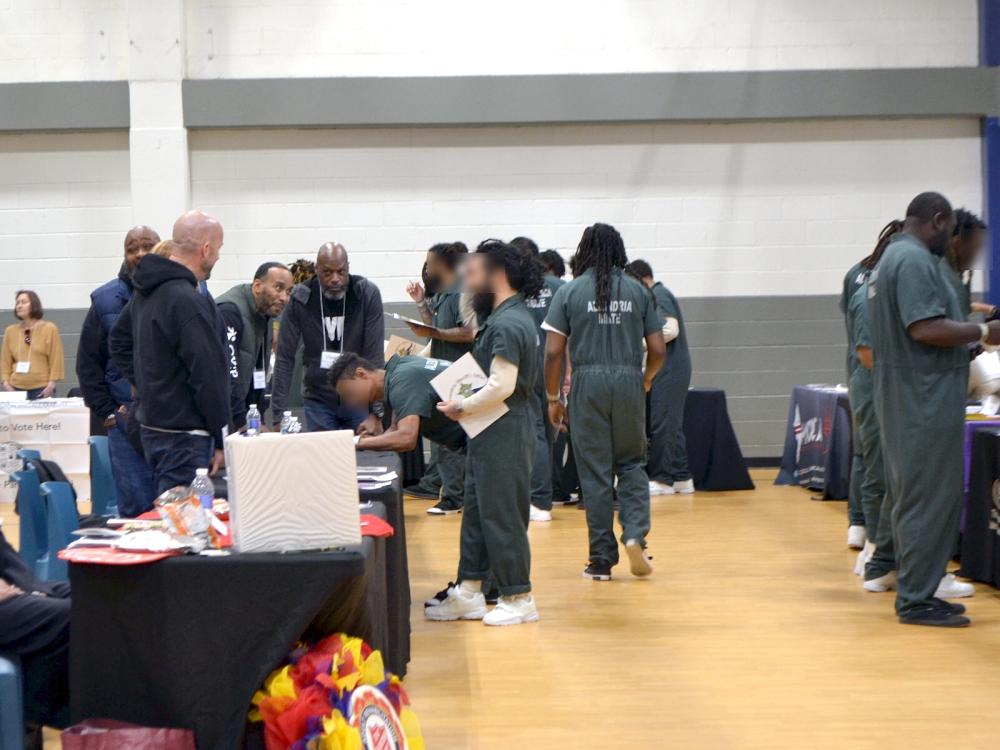 Several inmates in green jumpsuits speaking with exhibitors at tables in a gym