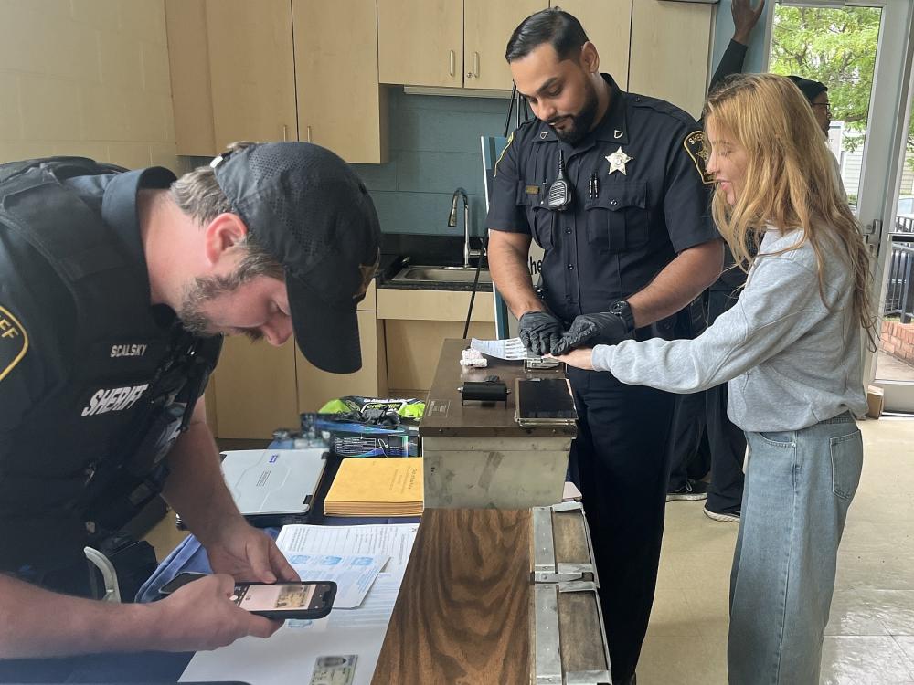 A civilian and two deputies in blue uniforms participating in an expungement clinic, one processing a photo ID and the other fingerprinting the civilian