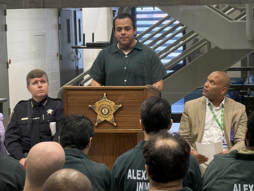 Inmate in a green jumpsuit speaking at a podium with a Sheriff's star on the front