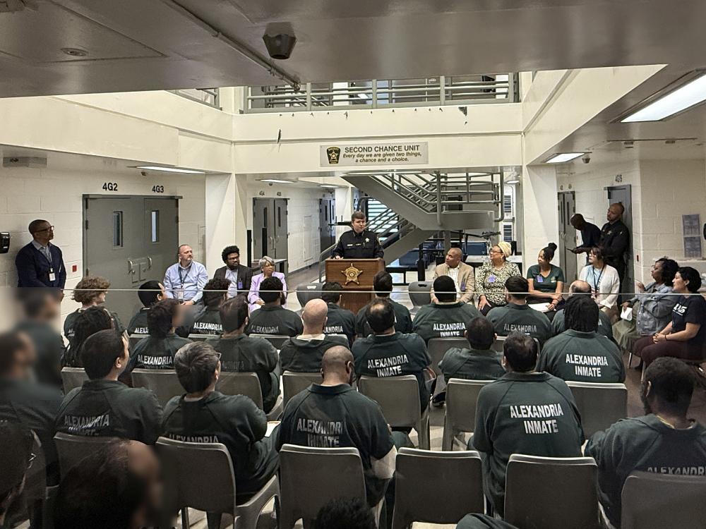 Sheriff standing at podium in front of several seated inmates wearing green jumpsuits and visible from the back