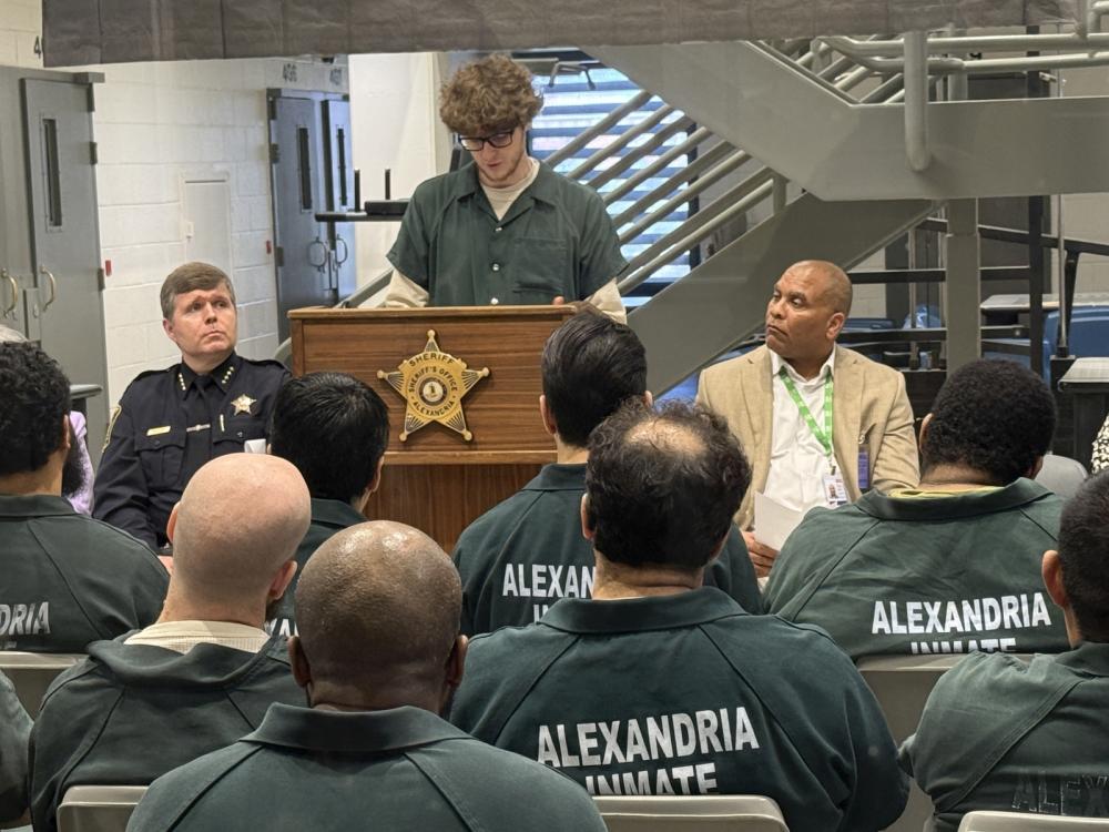 Inmate in a green jumpsuit speaking at a podium with a Sheriff's star on the front