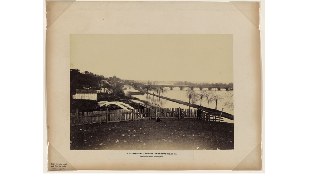 Photograph showing three men in Georgetown in the foreground with the Aqueduct Bridge and C&O Canal in the background.