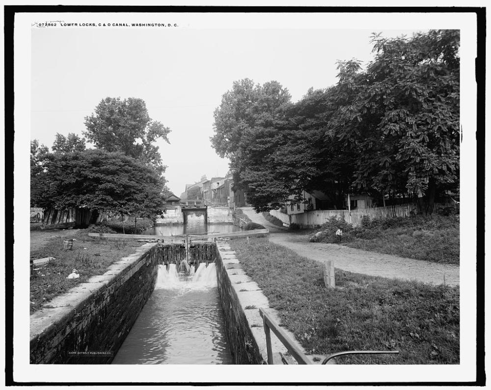 A black and white photo shows a narrow lock with gates in the background allowing water to enter from the larger basin beyond.