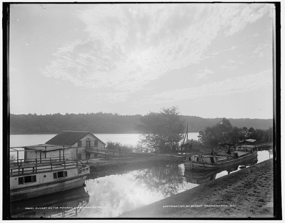 Black and white photo of two canal boats in a canal paralleling the Potomac River. A towpath is visible in the foreground.