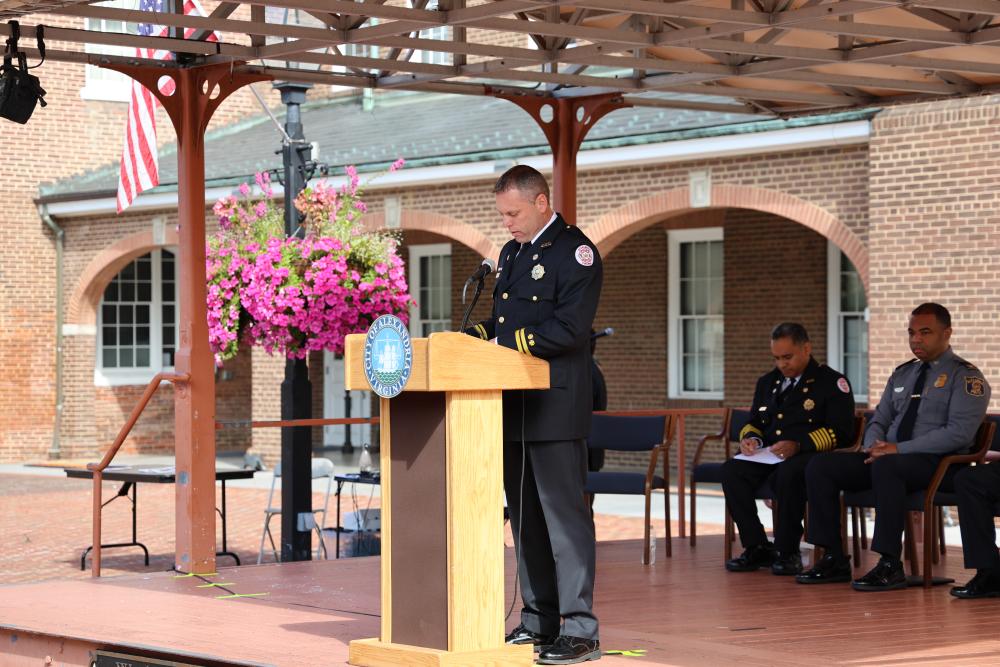 A person in firefighter uniform is giving a speech at a podium decorated with flowers, under a wooden shelter, at a formal event. Flags are visible in the background, and two individuals in uniform are seated nearby.