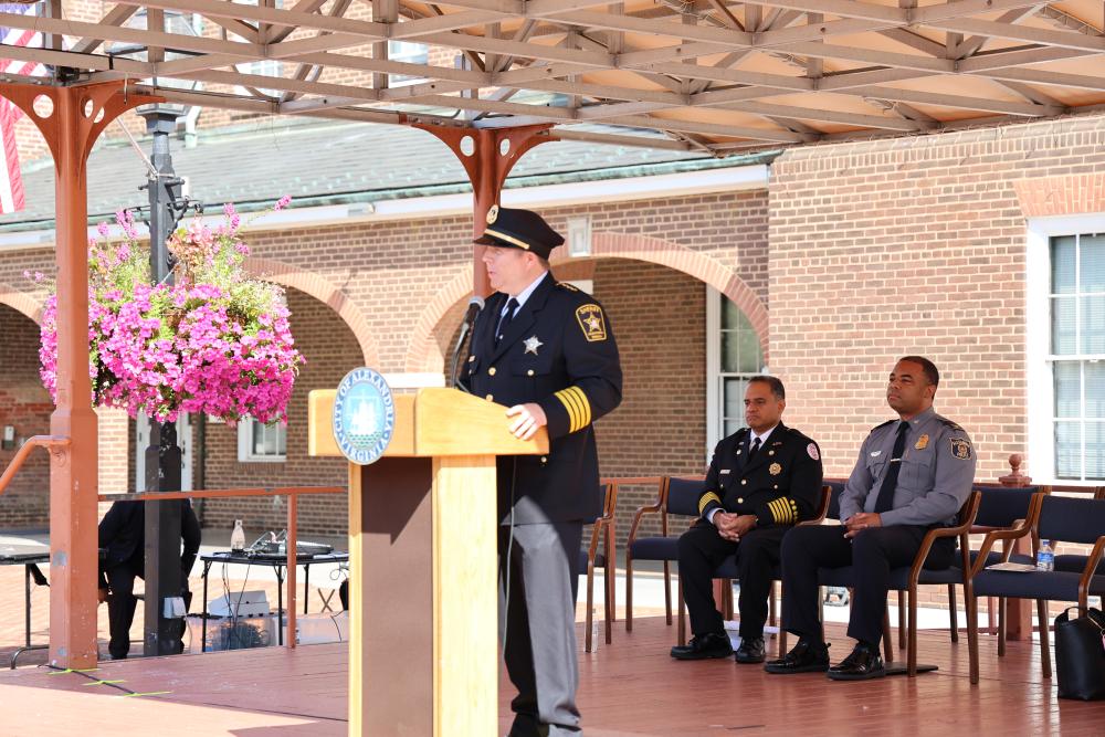 The sheriff in formal uniform is delivering a speech at a podium under a wooden pergola. Two other officers are seated nearby, listening attentively. Their backdrop is a brick building with a pleasant floral decoration.