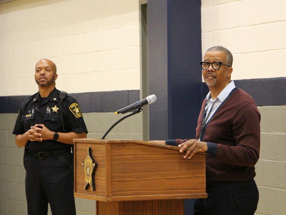Deputy in a blue uniform standing next to a civilan employee who is speaking at a podium