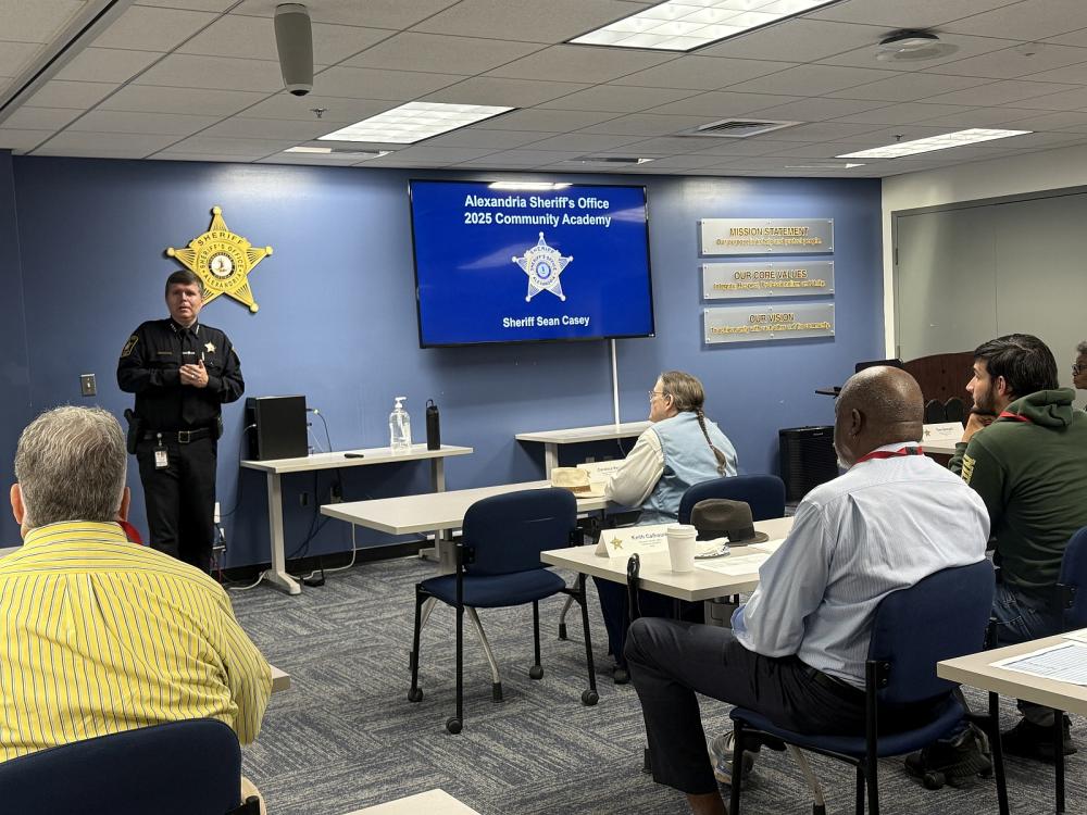 Sheriff in blue uniform speaking to community members seated at tables in the roll call room