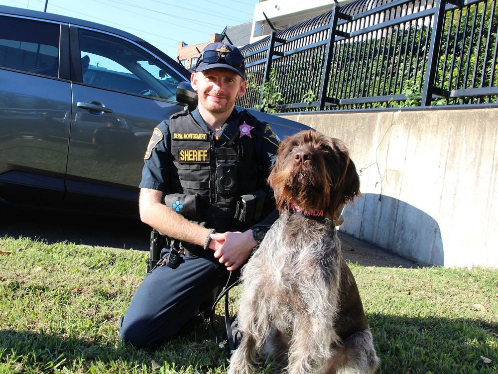Deputy in a blue uniform wearing a pink badge and his K9 partner, a German wiredhaired pointer wearing a black collar with pink letters reading K9 Aida 