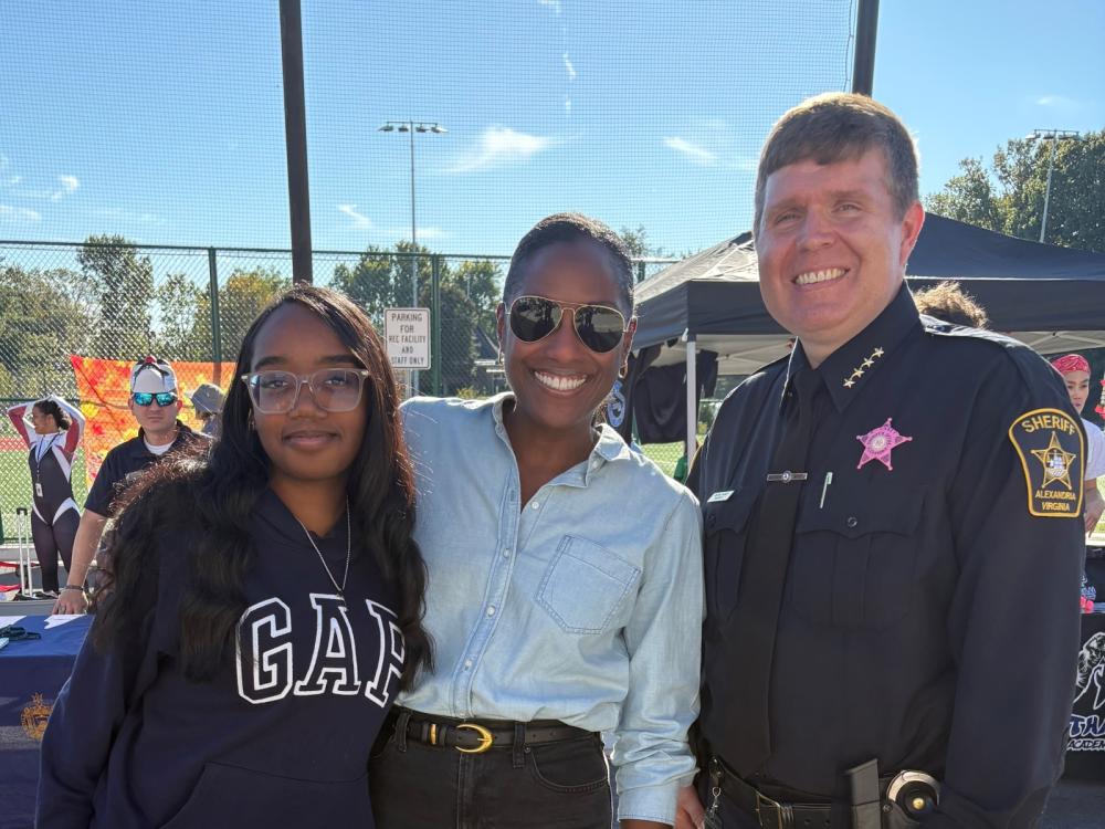 Sheriff in blue uniform wearing a pink badge and standing next two community members at an outdoor event