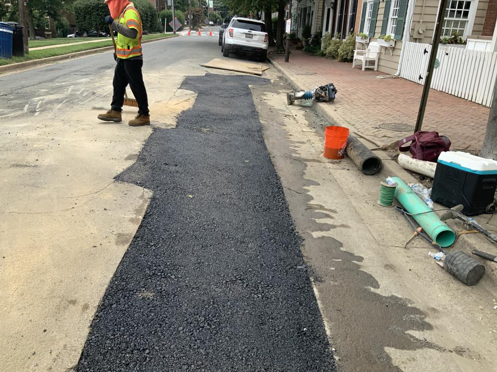 A worker stands next to a strip of fresh asphalt in the road. A turquoise and black sewer pipe awaiting installation are set next to the curb.