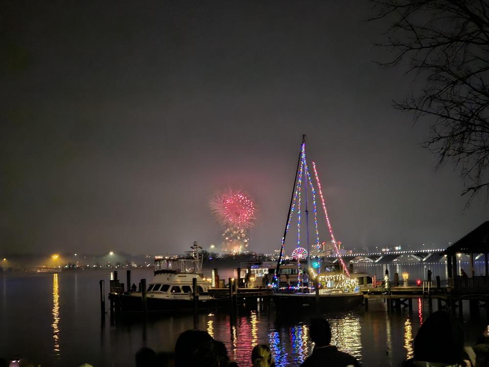 Fireworks in the distance behind a sailboat with holiday lights strung along its rigging.