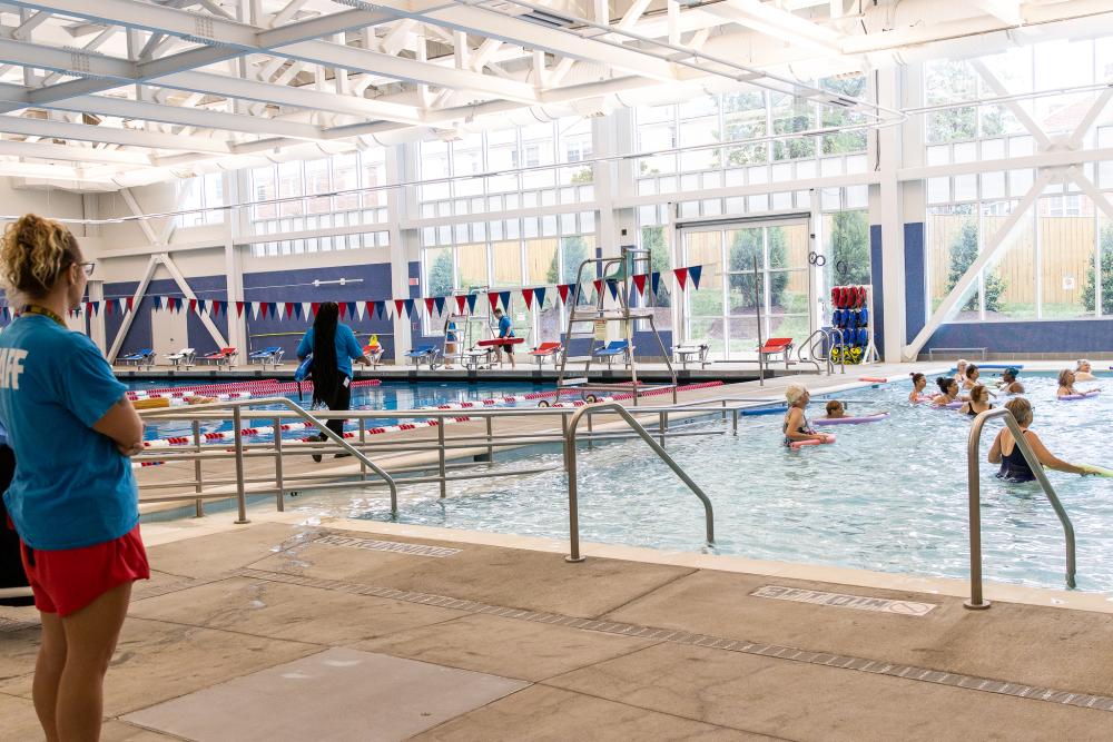 Minnie Howard's therapeutic pool featuring a group of community members engaged in an aquatics program, while a lifeguard stands on the left side facing them, another lifeguard walks between the two pools, and a third lifeguard sitting on his lifeguard chair