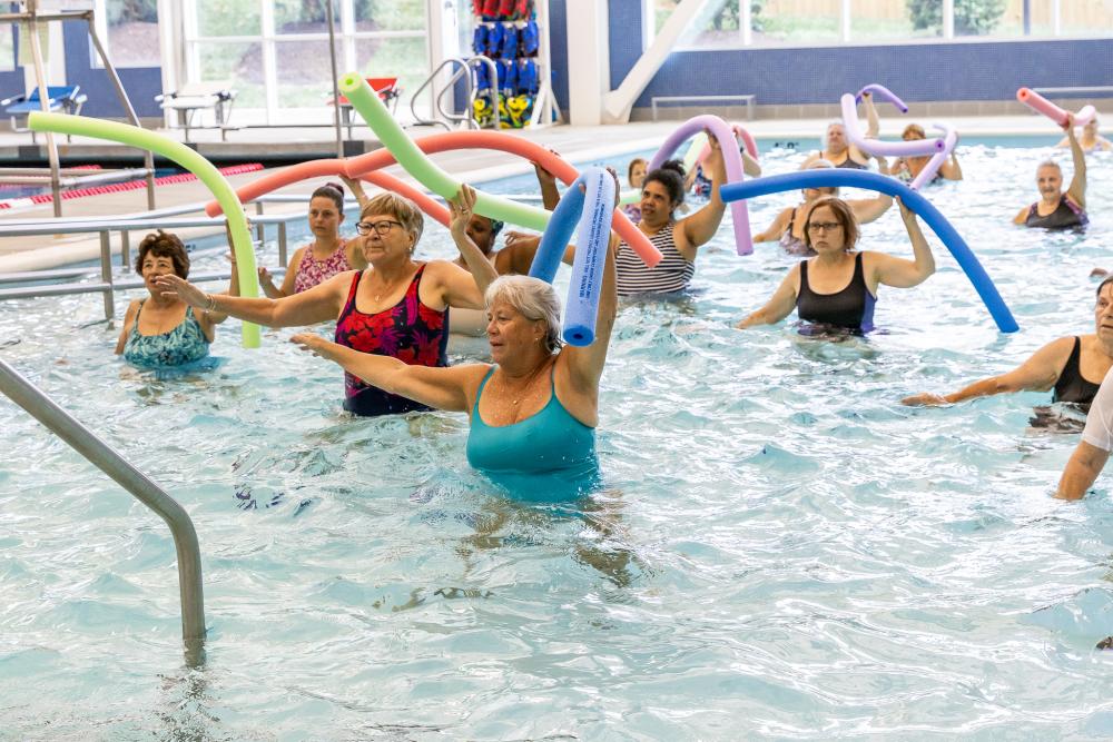 Low Impact Exercise in the therapeutic pool at Minnie Howard with several participants smiling and engaging with pool noodles in one hand and the other stretched out to the side