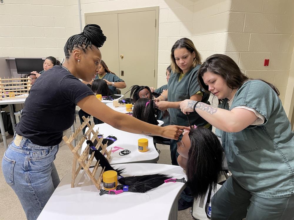 Hair braiding instructor providing a student-inmate with guidance on braiding hair on a mannequin head with four other student-inmates looking and working on braiding their mannequin heads