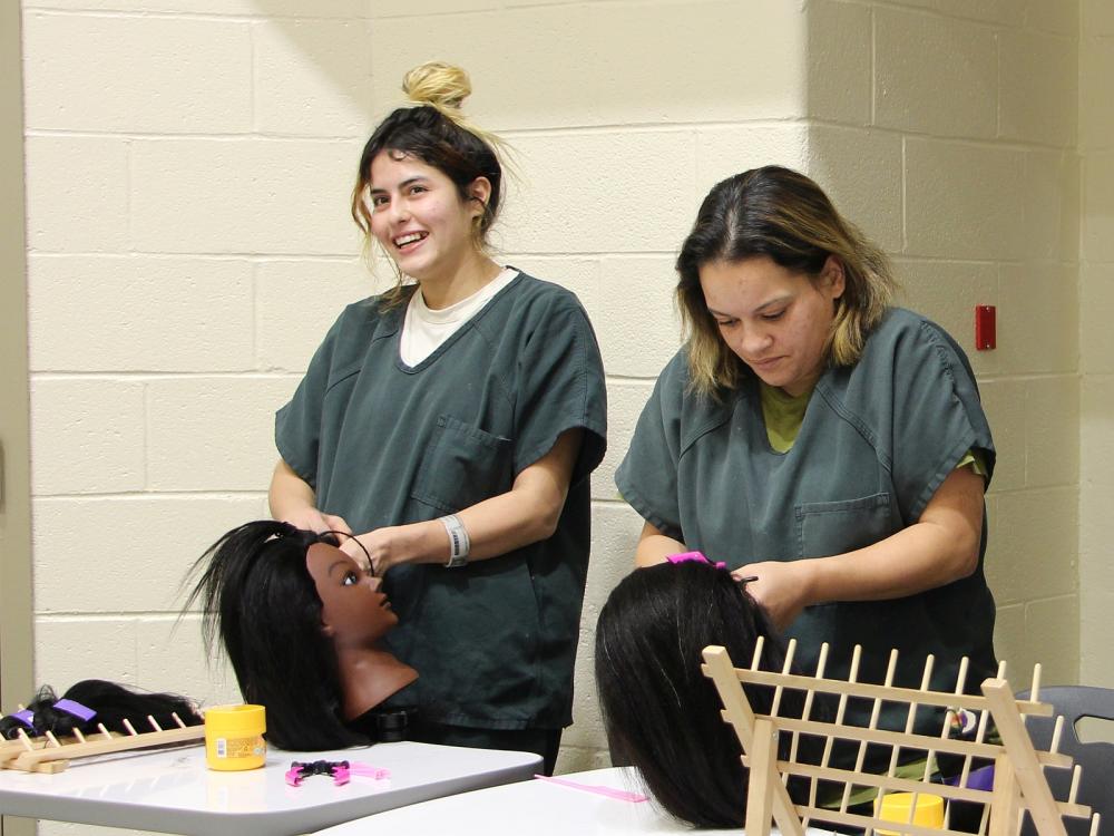 Two inmates in green uniforms braiding the hair on two mannequin heads