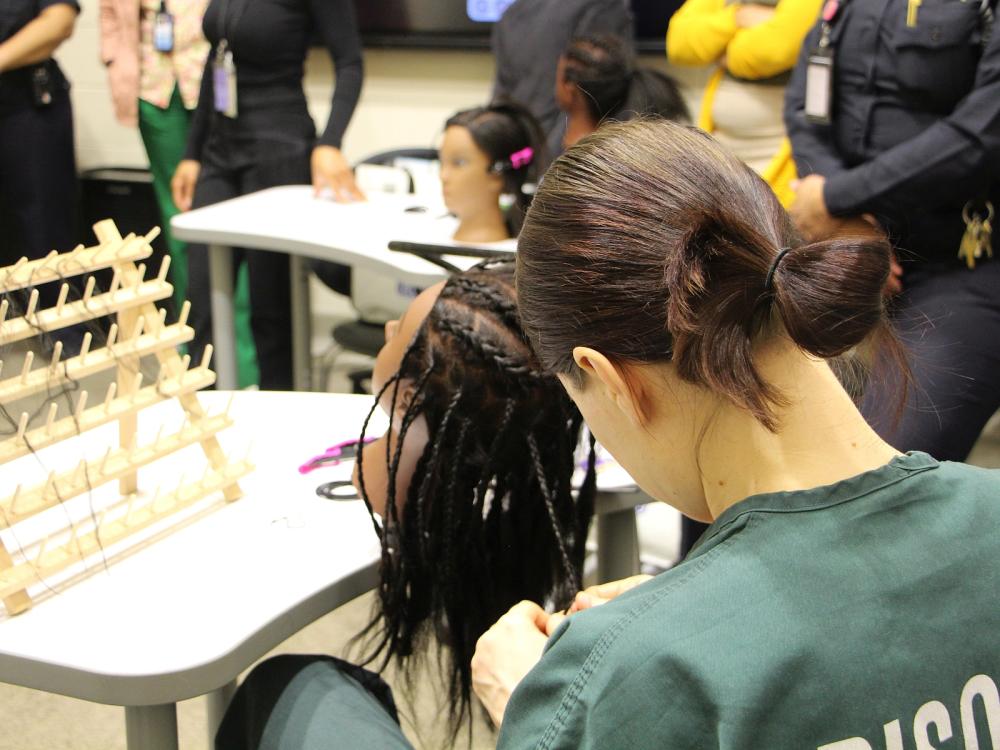 View from behind of an student wearing a green prisoner shirt and braiding hair on a mannequin head