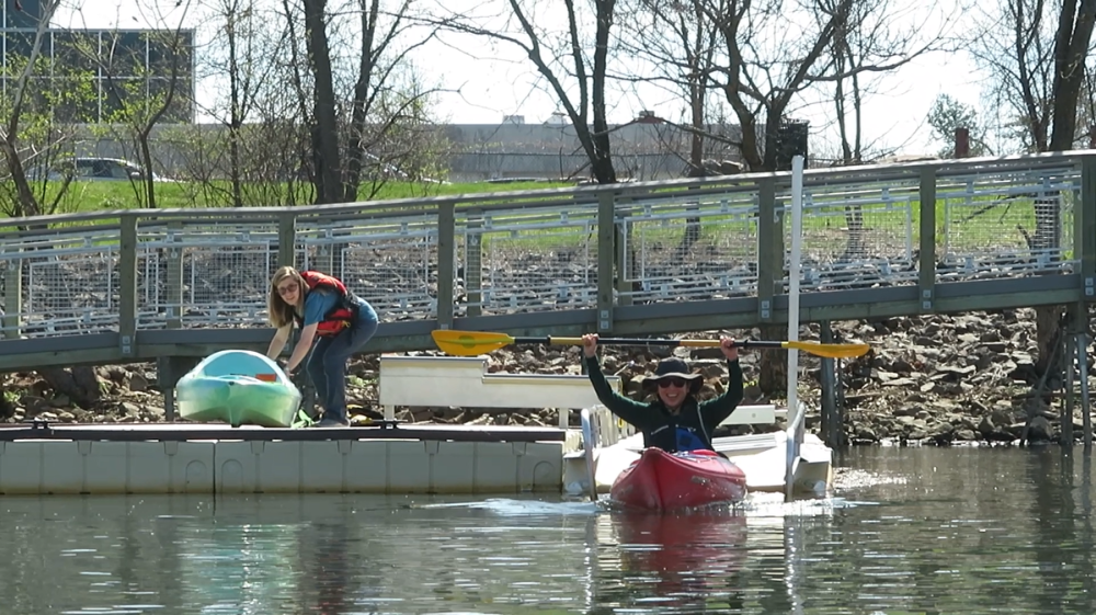 Woman on the deck while holding onto a kayak; another woman in a kayak on the launch with both arms up holding a paddle and smiling wide
