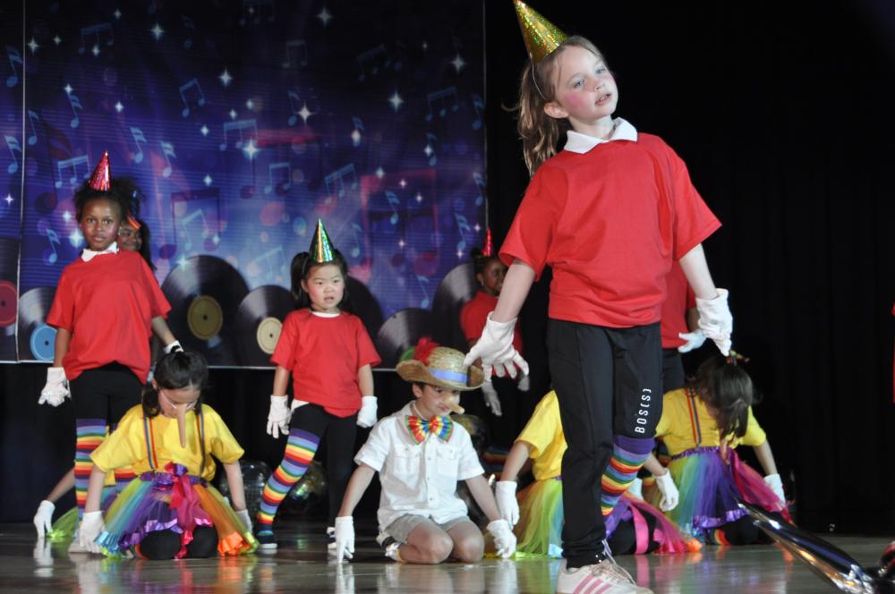 Kids dressed like puppets kneeling down on stage while kids wearing red shirts with party hats are standing up