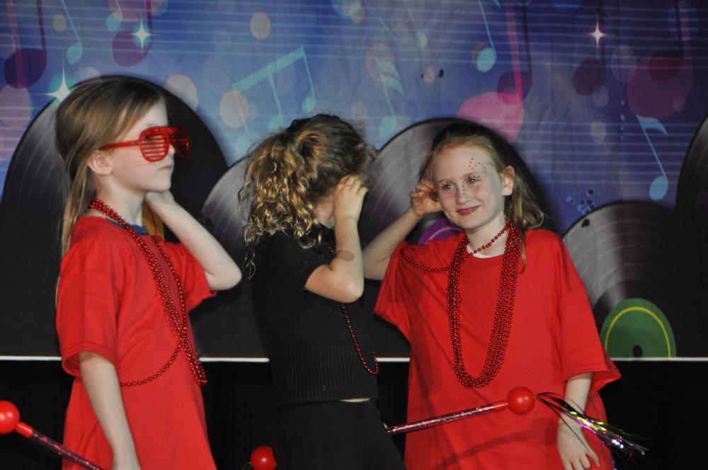 Three girls on stage with 2 girls wearing red clothing and one girl in the middle wearing black