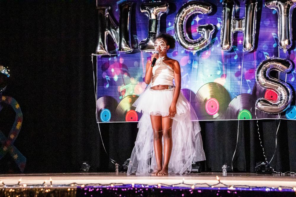 Performer in a white tulle dress with a white painted face and holding the mic