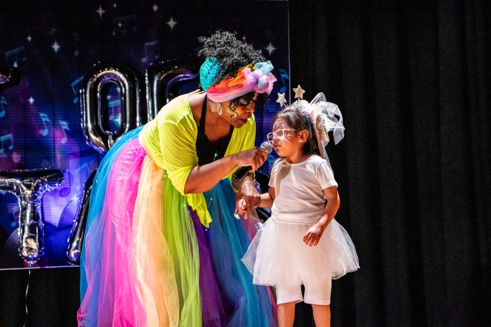 Emcee in a colorful dress and head wrap holding a mic for a little girl dressed in white