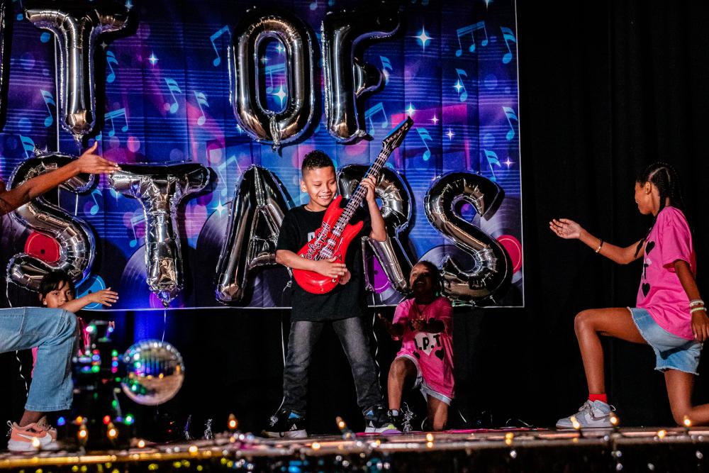 Performer in the middle holding a red, blow up guitar, while other performers are kneeling down around him