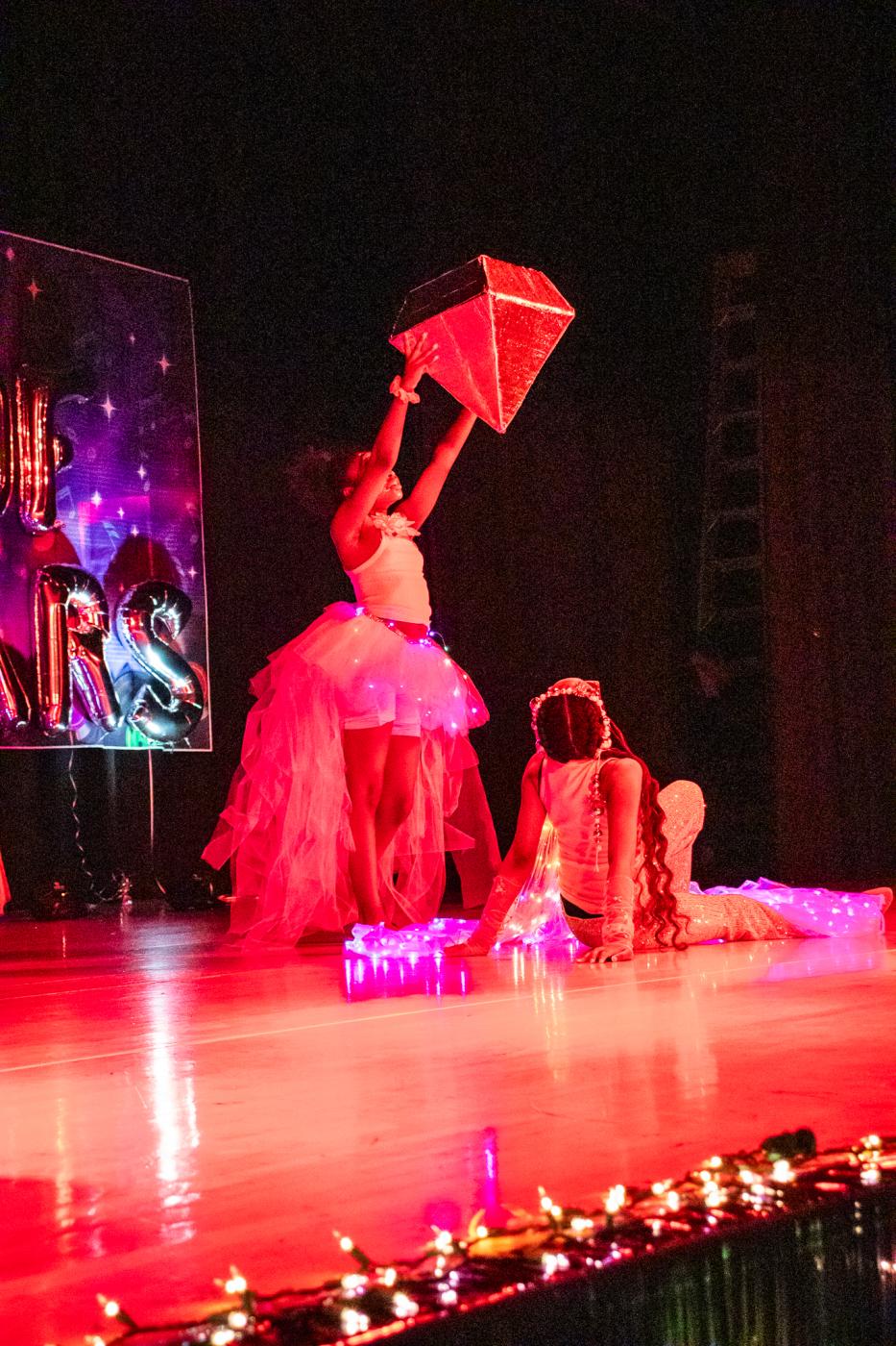 Performer in a dress standing while holding a giant diamond with another performer posing on the floor. Red light shining on both of them