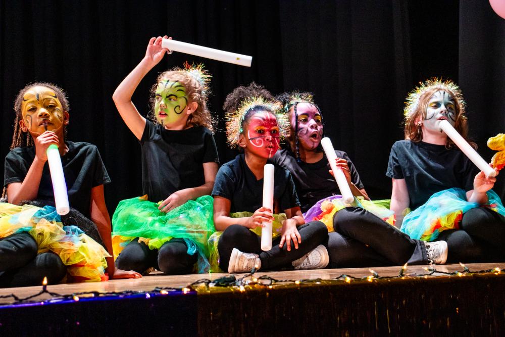 Child performers sitting on the stage dressed in black with colorful painted faces while holding white tubes