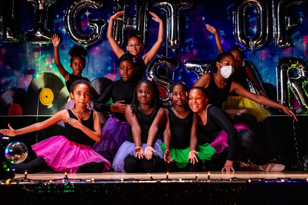 Performers in black sitting on the stage with colorful tutus and posing