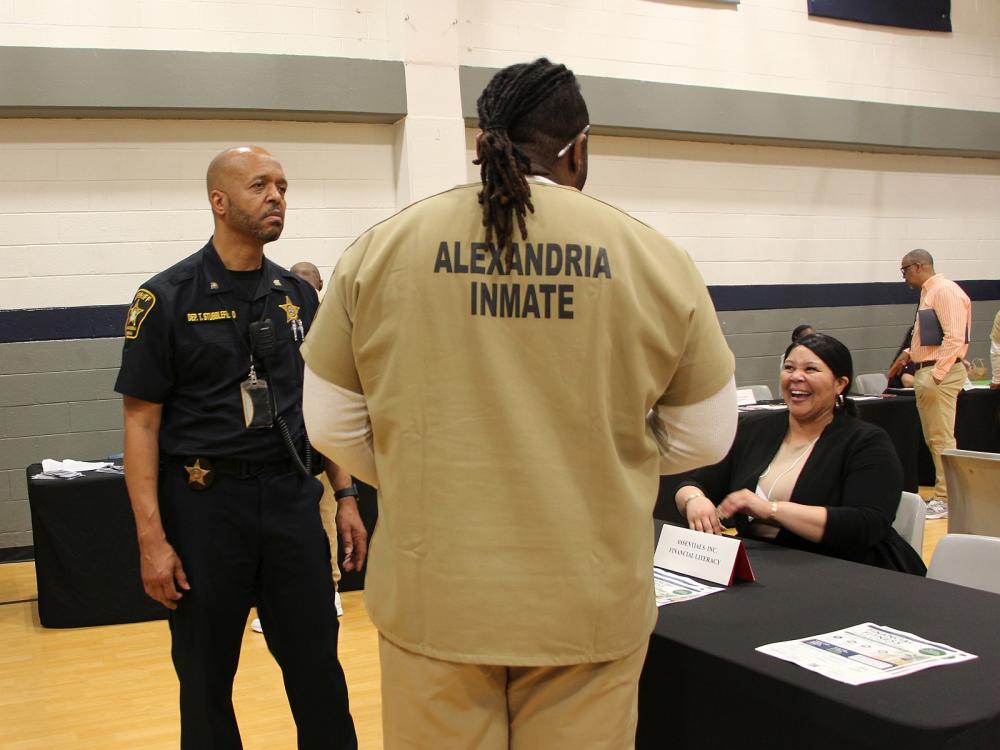Deputy in a blue uniform, inmate wearing a tan uniform that says Alexandria inmate on the back, and a business representative interacting with each other at a reentry resource fair