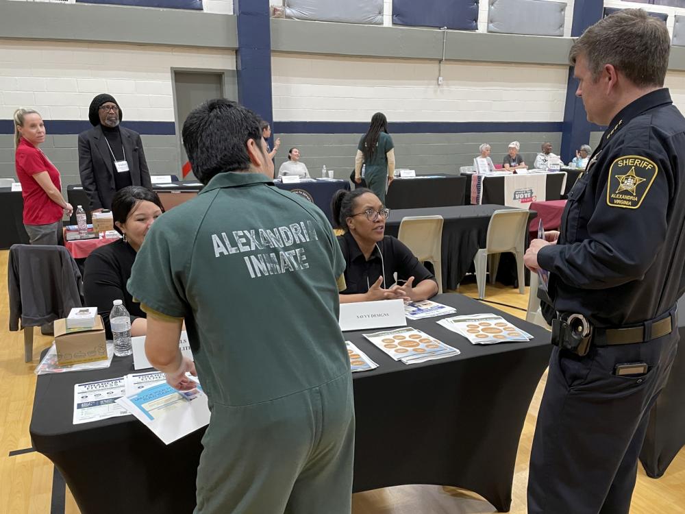 Inmate in green uniform that says Prisoner on the back, Sheriff in blue uniform and two business representatives interacting with each at the Reentry Resource Fair