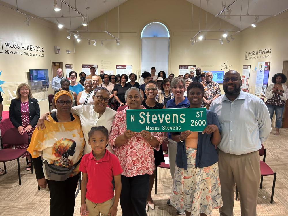 A group of people stands together indoors, holding a street sign labeled "N Stevens St - Moses Stevens." They appear to be in a museum or exhibit space with informational displays in the background, including one about Moss H. Kendrix. Chairs are arranged in rows, and some attendees are seated, smiling at the camera.