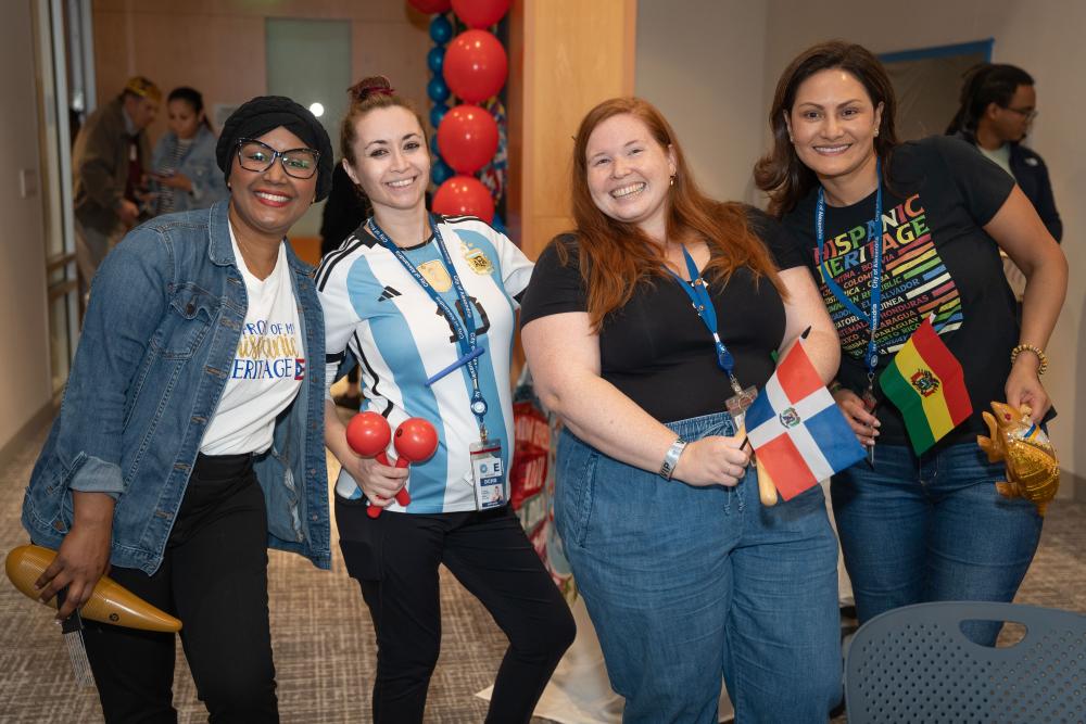 four women, two with flags and one with maracas standing and smiling at camera