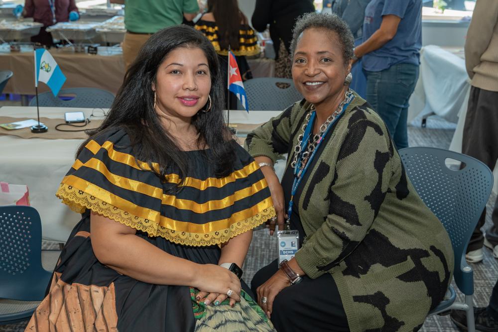woman with black and yellow dress and woman with green and black sweater sit next to each other and smile at camera