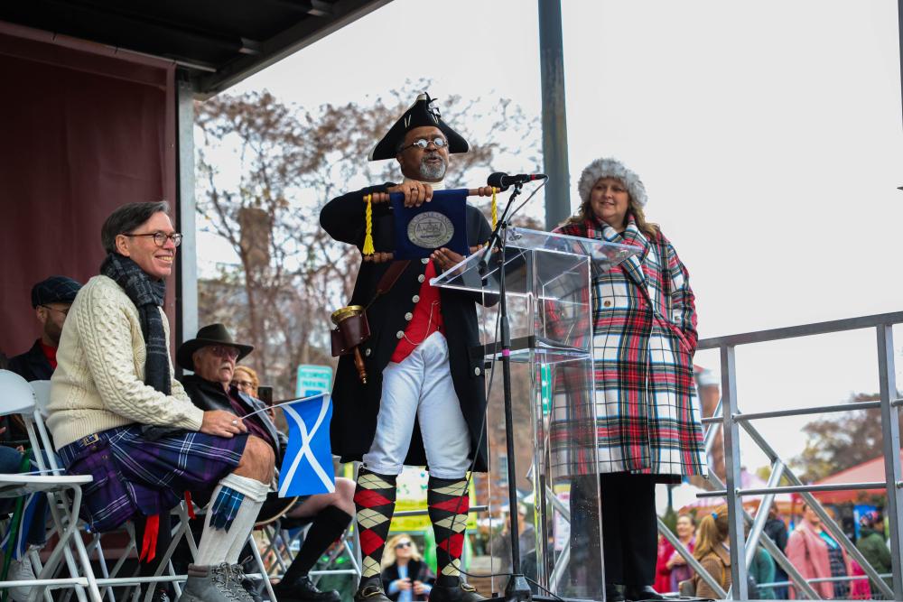 town crier on stage with a scroll surrounded by two people, one on the left sitting and one on the right standing