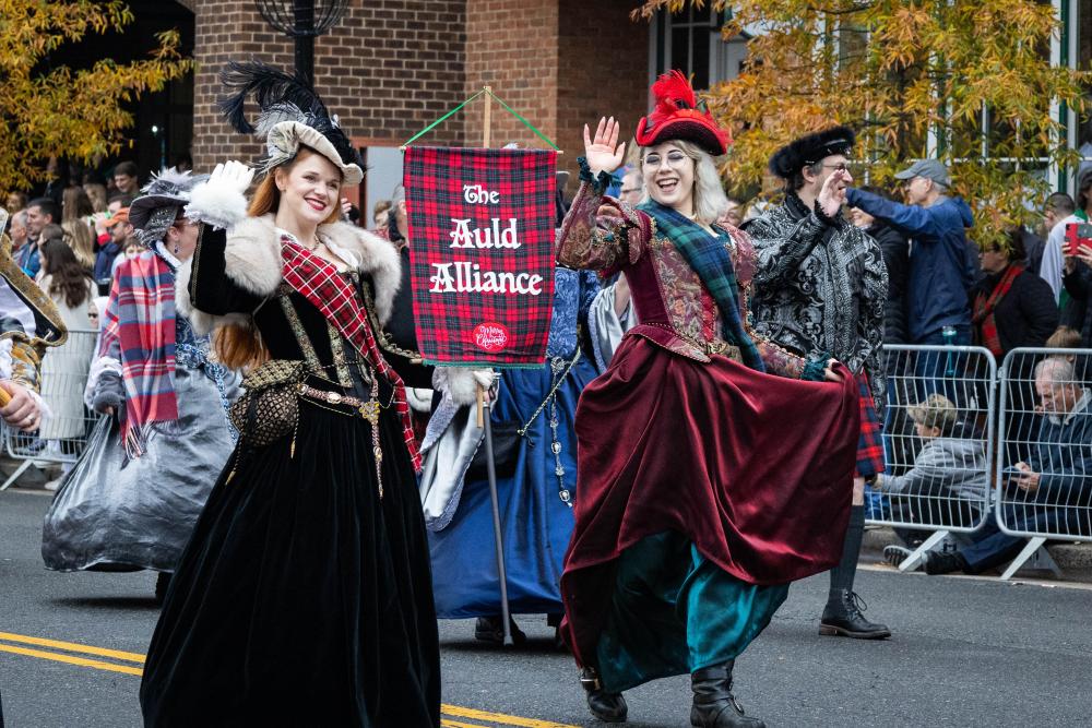 two women in elaborate dresses in hats walking in the parade holding a banner that says the Auld Alliance
